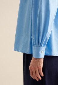 Himmelblaues Langarmhemd mit knöpfbarem Bündchen und Raffungen an den Ärmeln. Aus glattem Baumwollmaterial mit einem lässigen Schnitt.