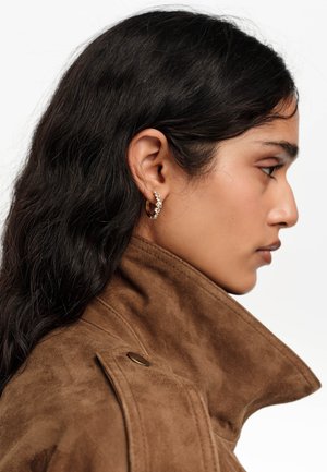 Młoda kobieta z długimi ciemnymi włosami, ubrana w brązową zamszową kurtkę i złotą kolczyk koło z przezroczystymi kamieniami szlachetnymi, zwrócona w prawo w widoku bocznym.
