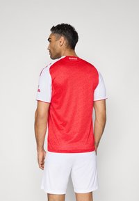 Rode sportieve trui met subtiel patroon, witte mouwen, ronde hals en bedrijfslogo op de achterkant, gedragen met witte shorts.