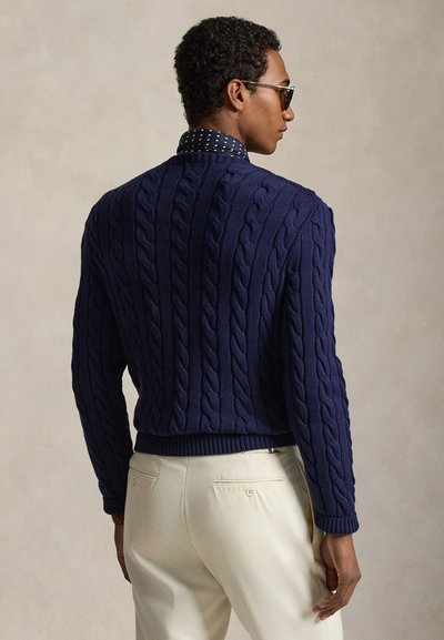 Άνδρας πλάγια φοράει γυαλιά ηλίου, μπλε σκούνο πουλόβερ με πλέξη καλώδιο, μπλε πουκάμισο με πουά και παντελόνι εκρού.