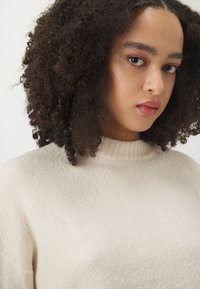 Cream-colored sweater with a ribbed high neckline, soft texture, and subtle sheen. Displays a relaxed fit and minimalistic design details.
