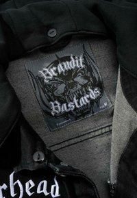 Svart tygjackas insida med en sydd etikett som har en skull-grafik. Etiketten visar "Brandit Bastards" och storlek "M." Knappar synliga.