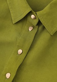 Material de culoare verde oliv cu o finisare texturată; prezintă un guler clasic și capse aurii pe lungimea plăcuței.