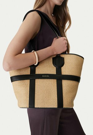 Kobieta ubrana w bezrękawnik i spodnie trzyma na ramieniu dużą beżową wiklinową torbę typu tote z czarnymi skórzanymi uchwytami i wykończeniami.