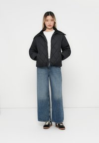 Giacca nera trapuntata con colletto alto, indossata sopra una maglietta bianca. Abbinata a jeans blu a gamba larga e sneakers nere. Design semplice e moderno.