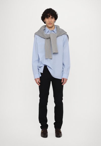 Helehedel sinine triibuline särk, hall soonik, mis on õlgadele tositud, mustad püksid ja tumedad kingad, millel on detailne aktsent. Ühtlane valge taust.