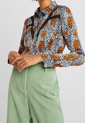 Femme portant un chemisier boutonné à manches longues avec un imprimé léopard orange et noir et un pantalon en velours côtelé vert clair taille haute.