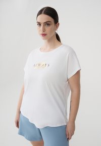 Donna con i capelli scuri raccolti in una coda di cavallo, che indossa una maglietta bianca con la scritta "ALWAYS" e pantaloni corti blu, in piedi contro uno sfondo chiaro e uniforme.