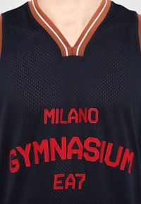 Basketbalový dres námořnické barvy s V-výstřihem, s červeným vyšívaným textem: "MILANO GYMNASIUM EA7." Síťovina s kontrastními hnědými akcenty na ramenou.