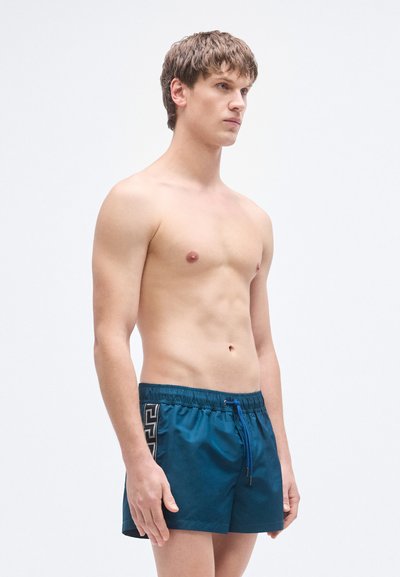 Μαγιό βερμούδα σε σκούρο τυρκουάζ χρώμα, με ελαστική μέση και μπλε κορδόνι, καθώς και αντίθετο μαύρο γεωμετρικό σχέδιο στο πλάι.