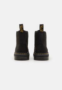 Botas de tobillo de ante negro con una textura suave, costuras amarillas a lo largo de la suela, y lengüetas con texto amarillo sobre un fondo negro.