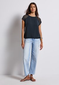 Zwarte top met korte mouwen en doorschijnende accenten, gecombineerd met lichtblauwe wijde jeans. De outfit wordt compleet gemaakt met platte sandalen met bandjes.