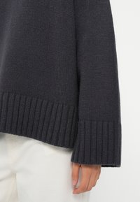 Dunkelgrauer gestrickter Pullover mit Rippbund und -ärmeln. Die Textur ist grob und der Schnitt ist leicht lässig. Hand auf hellem Hintergrund sichtbar.