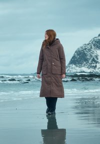 Bruine, lange pufferjas met een capuchon, twee voorkruiszakken en een gladde textuur, gedragen over zwarte broeken op een strand met een rotsachtige kust.