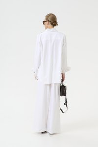 Vit oversized skjorta med långa ärmar, matchad med vida vita byxor. Svart handväska hängande över axeln, minimalistisk design.