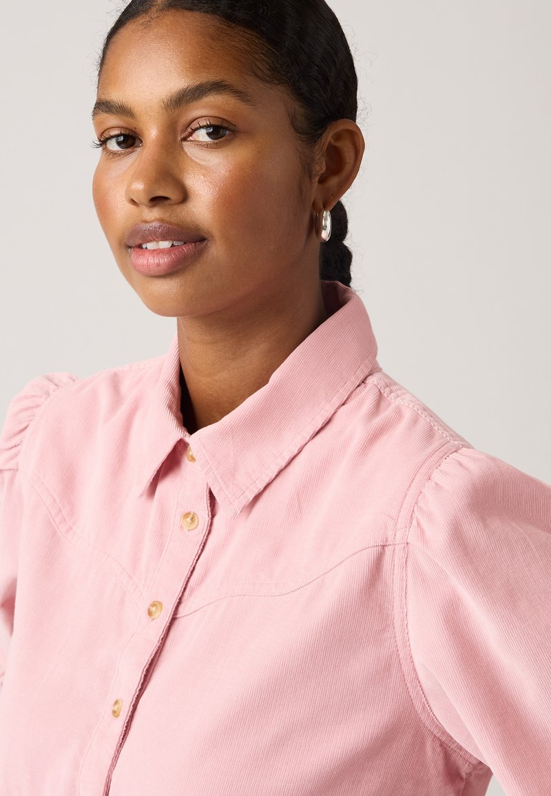 Chemise en velours côtelé rose avec un col boutonné, des manches bouffantes et des détails de couture visibles. Présente des boutons clairs et un tissu texturé.