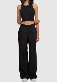 Personne portant un crop top noir sans manches, un pantalon noir taille haute à jambes larges et des baskets blanches, debout devant un fond uni.