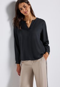 Donna che indossa una blusa nera a maniche lunghe con scollo a V e pantaloni beige con striscia nera, in piedi contro un muro bianco con una mano in tasca.