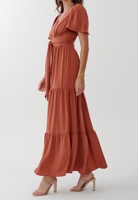 Maxiklänning i rostfärg med korta puffärmar, en åtdragen midja och flerlagad kjol. Tyget har en strukturerad yta. Bärs med beige blockklacksandaler.