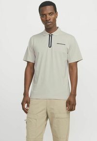 Lys grå polo skjorte med en sort lynlås og en diskret detalje på brystet; kombineret med beige cargo bukser, der har flere lommer.