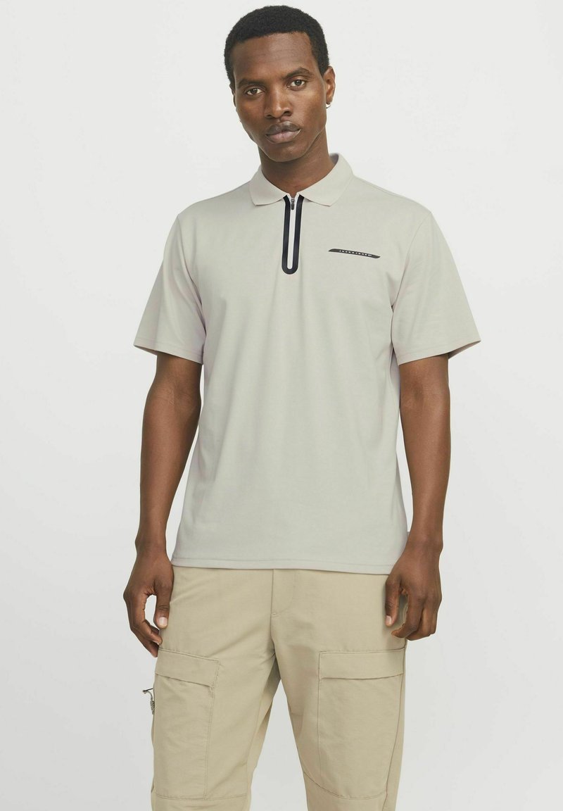 Lichtgrijs poloshirt met een zwarte rits en subtiele accenten op de borst; gecombineerd met beige cargobroek met meerdere zakken.