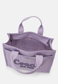 Lilanvärinen kangaslaukku, jossa on kaksi kantokahvaa, vetoketjusuljin ja etutasku. Laukussa on kirjailtu "Coach New York" -logo ja sisällä etikettitieto.