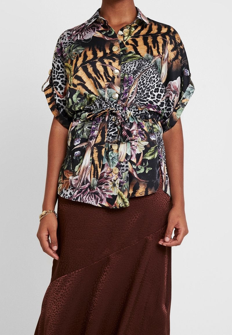 Chemise à manches courtes, à boutons, avec un imprimé animal et floral vibrant, présentant des nuances de noir, orange et vert. Détail de nœud à la taille.