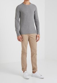 Graues Langarmshirt aus weichem Stoff, kombiniert mit beigen Chinos. Das Modell trägt weiße Sneakers und steht vor einem weißen Hintergrund.