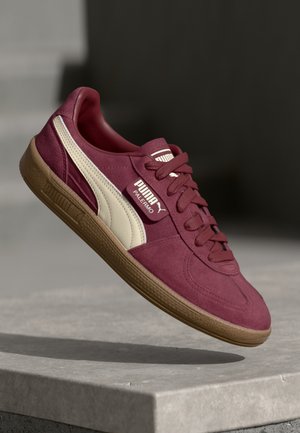 Baskets Puma Palermo bordeaux avec bande blanche et semelle en gomme flottant au-dessus d’une surface en béton sur fond sombre.