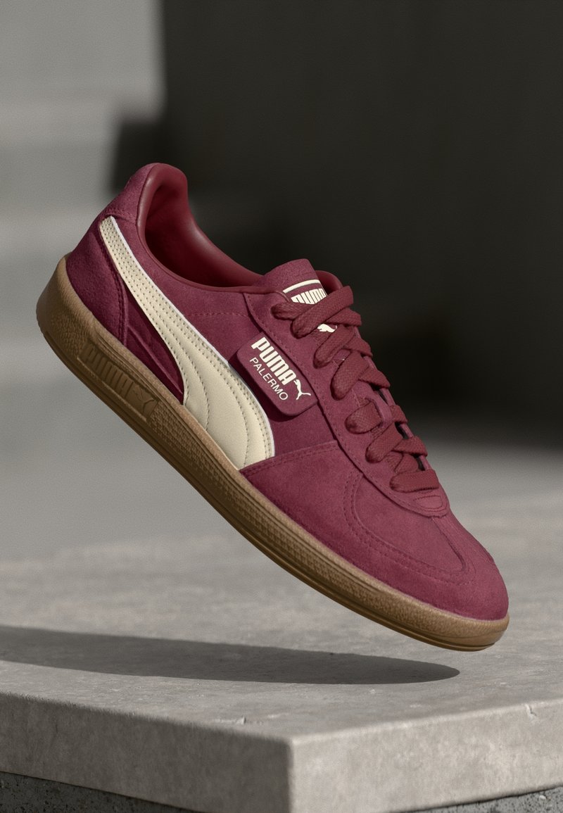 Baskets Puma Palermo bordeaux avec bande blanche et semelle en gomme flottant au-dessus d’une surface en béton sur fond sombre.