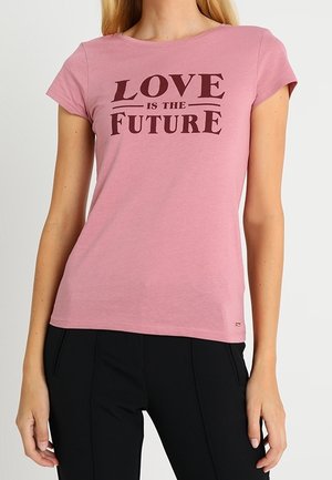 Frau trägt ein pinkes T-Shirt mit dem Text "LOVE IS THE FUTURE" in Dunkelrot, kombiniert mit einer schwarzen Hose, vor einem einfarbigen Hintergrund.