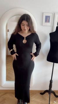 Donna che sistema un lungo vestito nero a coste con scollo a goccia, in piedi vicino a uno specchio a figura intera e a un manichino in una stanza con quadri incorniciati.