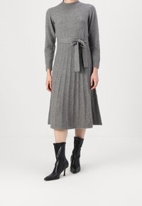 Robe grise en maille avec manches longues et taille cintrée, dotée d'une jupe plissée et d'une ceinture en tissu nouée. Portée avec des bottines noires.