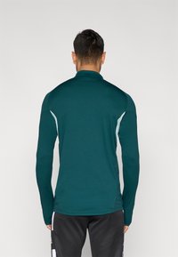 Langærmet sportstop i mørk teal, med polokrave og kontrasterende lyseblå detaljer på skuldre og sider. Glat stof.