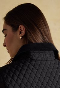 Femme aux cheveux bruns lisses porte de petites boucles d'oreilles en or en forme de créole et une veste matelassée noire avec "JOULES" brodé sur le col.