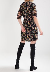 Kvinna iklädd en svart blommig klänning med rosa och gröna fåglar, kombinerad med svarta stövlar med klack som går över knäet, vänd bort med en hand bakom ryggen.