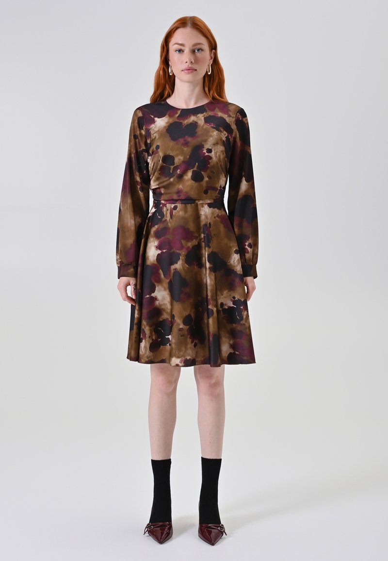 Een jurk met een bruine en paarse bloemenprint, lange mouwen, een aansluitende corsage en een uitlopende rok; gedragen met zwarte sokken en puntige schoenen.