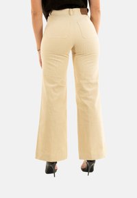Pantaloni a gamba larga in corduroy beige con vita alta, due tasche posteriori e una patch in pelle marrone sulla vita, abbinati a tacchi alti neri.