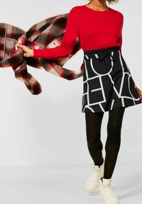 Kvinna i röd långärmad topp, svart och vit mönstrad kjol, svarta strumpbyxor och vita sneakers som håller en flygande röd och svart rutig jacka vid sidan av.