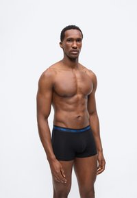 Μυώδης άνδρας που στέκεται φορώντας μαύρα μποξεράκια με μπλε ζώνη μέσης με τη μάρκα «Hugo» σε απλό λευκό φόντο.