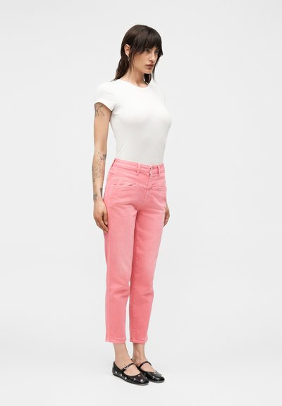 Γυναίκα με σκούρα μαλλιά και τατουάζ, που φοράει λευκό μπλουζάκι κοντομάνικο, ροζ τζιν ψηλόμεσα και μαύρα παπούτσια Mary Jane, στέκεται μπροστά σε απλό φόντο.