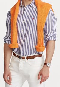 Herroutfit med en lila och vit randig skjorta med knappar, en orange kabelstickad tröja lagt över axlarna, vita byxor och ett brunt bälte.