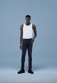 Top fără mâneci, alb, blugi skinny închiși la culoare și cizme negre cu șireturi. Modelul stă în fața unui fundal bleu deschis, evidențiind un stil minimalist.