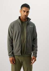 Jachetă din fleece gri cu guler înalt, închidere cu fermoar și logo pe partea stângă a pieptului; purtată peste o cămașă verde și asortată cu pantaloni corespunzători.