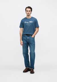 Μπλε βαμβακερό T-shirt με λευκό λογότυπο "Tommy Hilfiger", συνδυασμένο με μπλε τζιν τζιν και καφέ δερμάτινα μποτάκια, σε απλό φόντο.