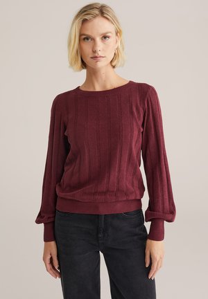 Burgunderfarbenes Strickpullover mit Rippstruktur, Rundhalsausschnitt und Ballonärmeln. Kombiniert mit dunklen Jeans für einen kontrastierenden Look.