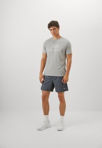 Light gray short-sleeve t-shirt with a white graphic logo, paired with dark gray shorts and white sneakers, displayed on a neutral background.