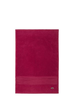 Serviette rectangulaire rouge foncé avec une bordure texturée près d’un bord et un petit logo crocodile vert dans un coin.