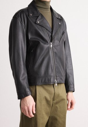 Personne portant un blouson de motard en cuir noir sur un col roulé olive et un pantalon vert, debout devant un fond clair et uni.