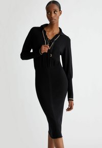 Zwarte gebreide jurk met een kraag, lange mouwen en een getailleerd silhouet. Opvallende details zijn een veterhals en gouden accentmanchetten.